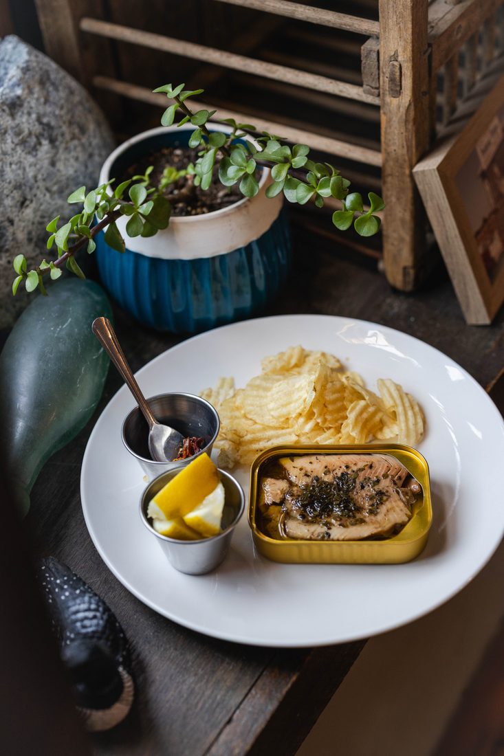 An open tin of fish plated with crinkle-cut chips, lemon, and chili crisp